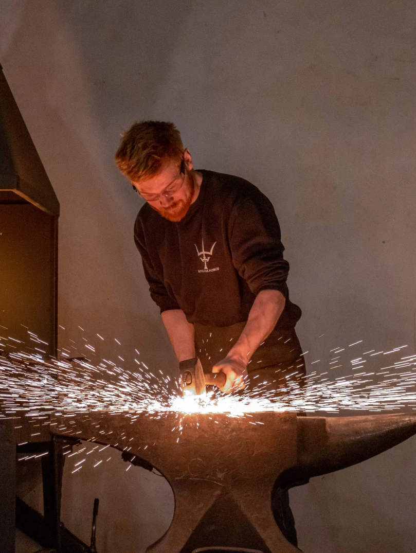 Blacksmith forging metal on an anvil with sparks flying and a furnace ablaze in the background