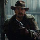 Close up still of Lee Scoresby (Lin-Manuel Miranda) from His Dark Materials (Series 2) (Bad Wolf). Lin-Manuel is wearing wearing a brown hat, brown jacket and is holding a metal handgun.