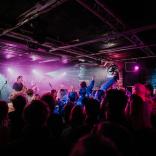 Person crowdsurfing in a dark room lit up by stage lights while a band plays on stage at Club Ifor Bach for the BBC 6 Music Festival