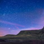 Outline of lake and mountain backdrop against a dark blue and purple night sky, dotted with blurry stars