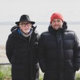Two peope wearing black bomber coats and hats (one orange beanie and one black wide brimmed) standing with their backs to the sea