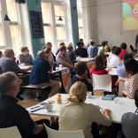 A large group of people sitting at white tables facing a projector screen as someone presents. The screen shows a background of colourful balloons with the word "Clwstwr" in white text in the middle.