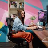 Medium shot of Robyn working at her wooden desk at Bomper Studios in Cardiff. Robyn is wearing an orange midi shirt and a blue shirt. The wall behind her is covered in blue, yellow and pink graphic patterns. 