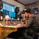 Jayce Lewis of Northstone Studios sits at a wooden mixing desk with monitors and large speakers in a dimly-lit studio.