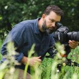 Close up shot of a person wearing a blue coloured shirt looking through a camera lens.