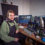 Person wearing dungarees over a green jumper with a check scarf sits at a desk with a laptop, monitor screen and microphone.