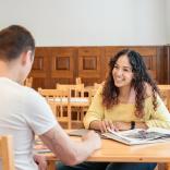 Close up shot of Yassmine from Bomper Studio talking to colleague. Yassmine is smiling, sitting at a wooden table wearing a yellow of the shoulder top.