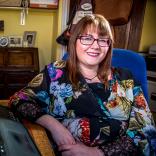 Medium shot of Rebecca Roberts. Rebecca is sitting at her desk at home in a yellow room. She's wearing a black dress with a colourful flower print, rectangle dark red glasses and has medium-length brown hair.