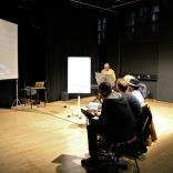 Group of people sit in a dimly lit hall surrounding by black walls. There is a person standing at the front talk. Next to them is a white paper flipboard. 