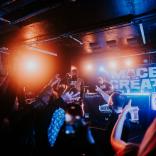 Photo of a dark gig venue. In the foreground, you see the audiences' hands in the air. On stage, Mace the Great performs under bright orange and blue lights. 