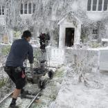 A person stands on a mini track holding a film camera. They are focusing on a white house covered in snow.