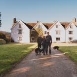 A man standing with a woman and a younger man in front of a large pink farm house on a sunny day