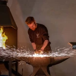 Blacksmith forging metal on an anvil with sparks flying and a furnace ablaze in the background