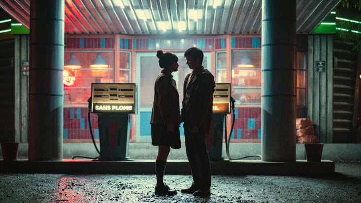 Shadow outline of Emma Mackey as Maeve Wiley and Asa Butterfield as Otis Milburn standing close together in front of a retro petrol station at night