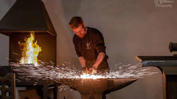Blacksmith forging metal on an anvil with sparks flying and a furnace ablaze in the background